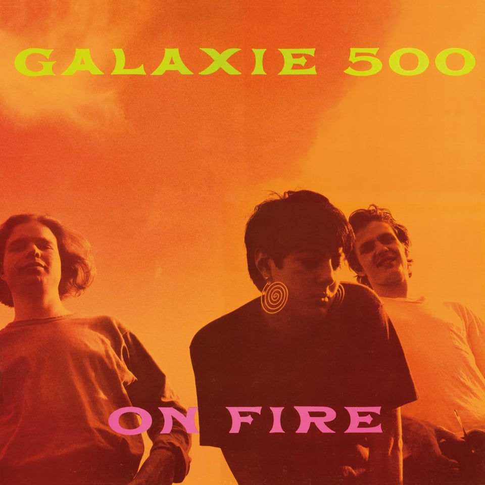 A photograph of the band, taken from a low angle on the ground looking up at them. They're dressed in T-shirts. There's a guy with a round face and longish light-colored hair, a woman in a dark shirt with short dark hair and large spiral earrings, and another guy with short moussed hair in a white t shirt. The whole thing is orange-tinted. A great deal of the picture is the sky above the three, and the sky is orange.