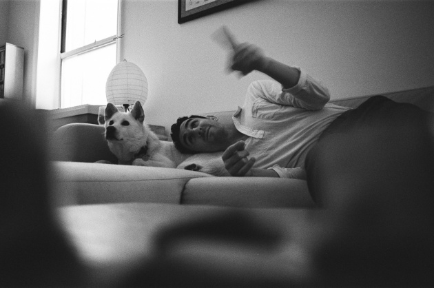 A man lies on a couch with his head on the midsection of a white dog.