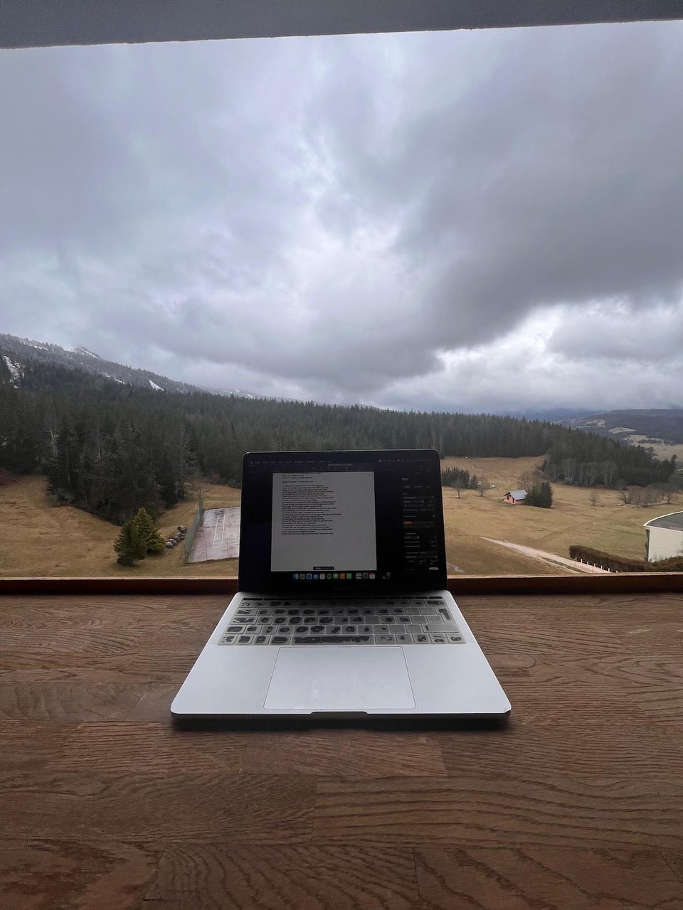 un ordinateur portable ouvert avec un traitement de texte en cours, posé sur la rambarde d'un balcon avec vue sur la montagne et le ciel gris