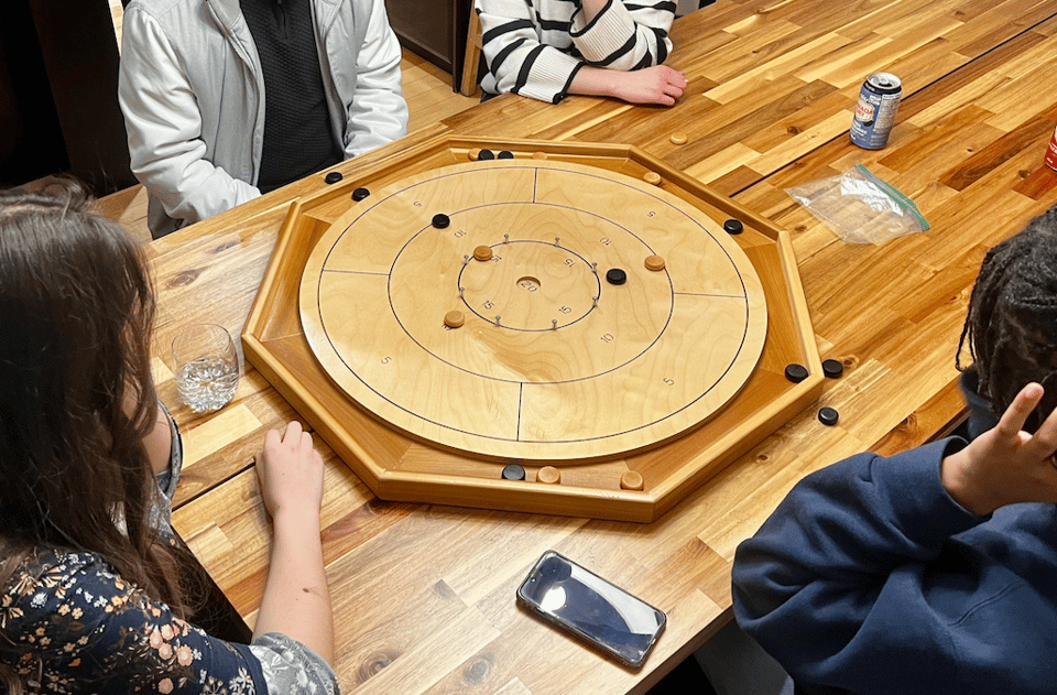 A photo from above of a Crokinole board on a table with 3 people around it playing the game.