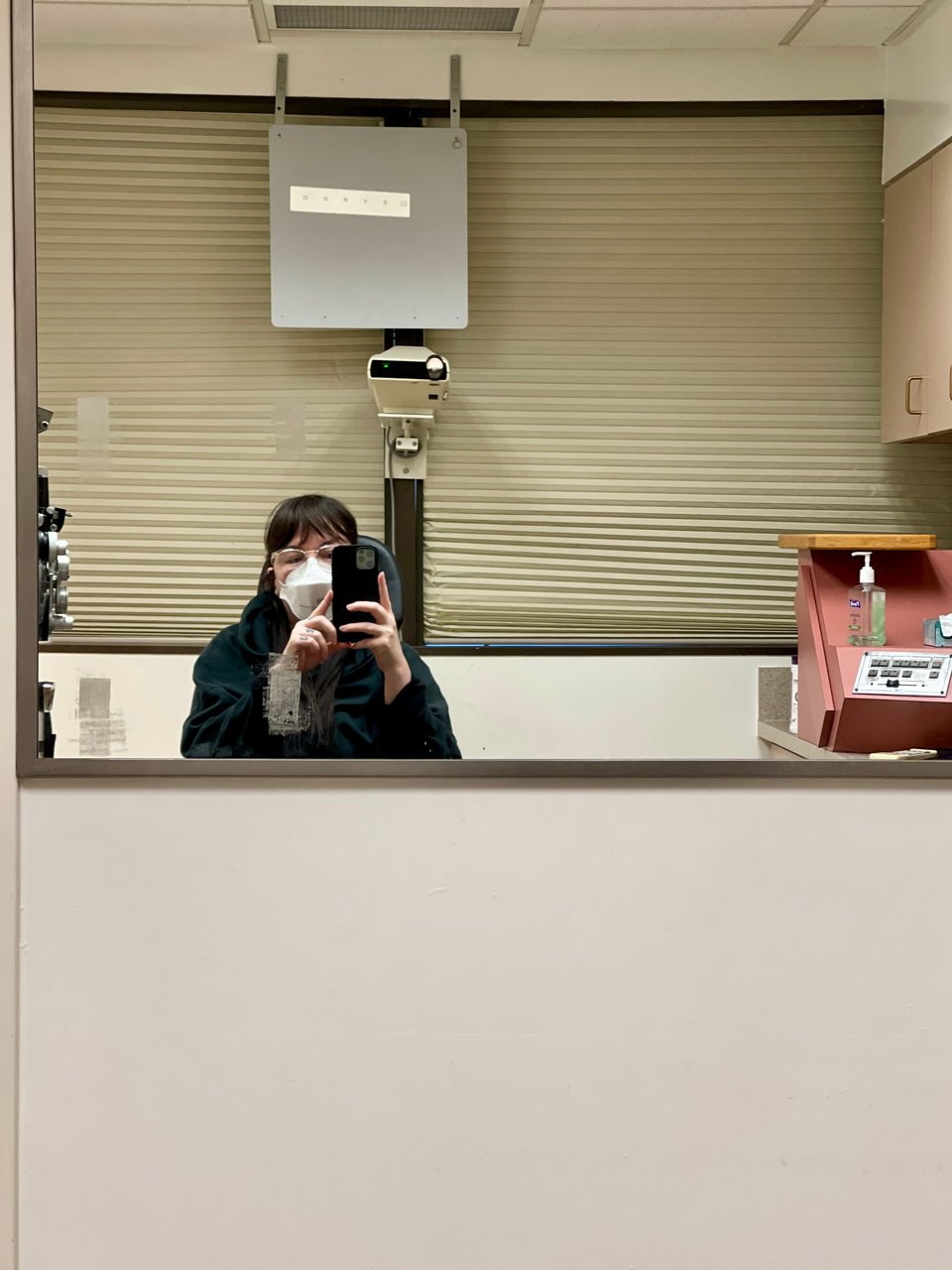 A masked person in patient room at a hospital taking a selfie in a mirror