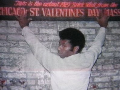 A man standing with his hands above his head against the wall. A sign says "This is the actual 1929 brick wall from the Chicago St Valentines Day masscare"