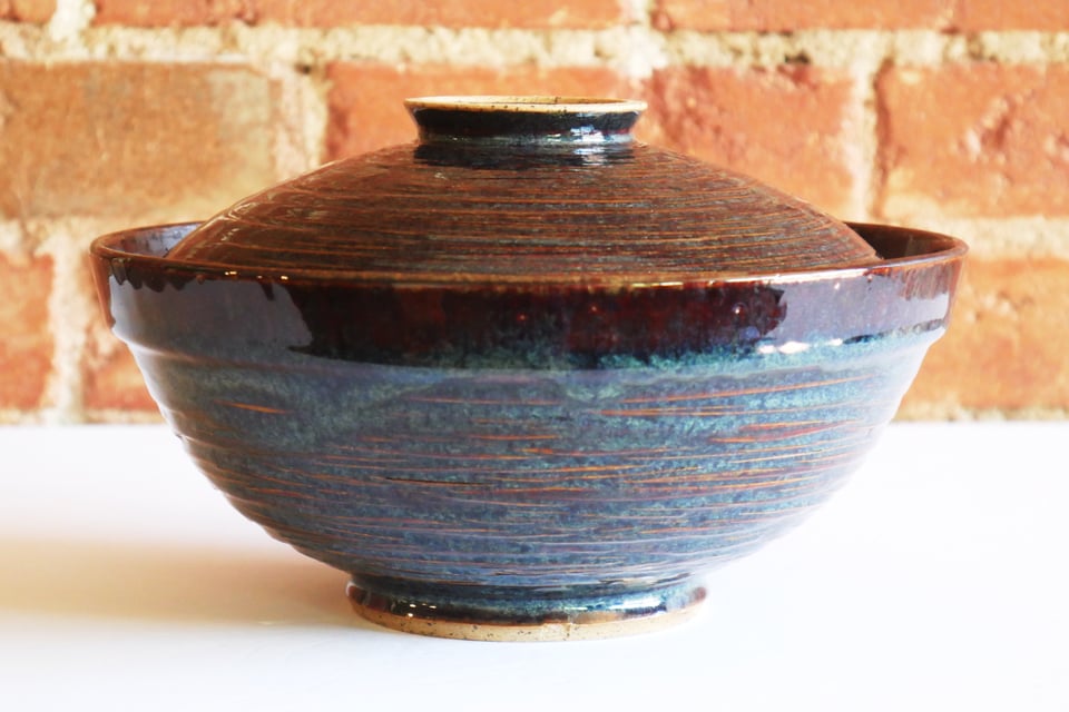 Large round-bottom ramen bowl with a wide rim and textured walls of glassy blues on browns. There's a deep foot ring at the base and a wide shallow lid that sits into the lip - also textured, and with a ring at the top for a handle.