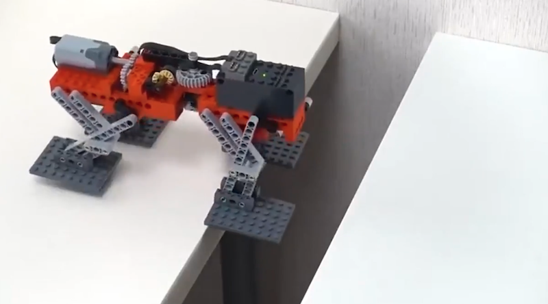 A gray-and-red four-legged robot, constructed out of Legos, is perched on the edge of a gap between two tabletops.