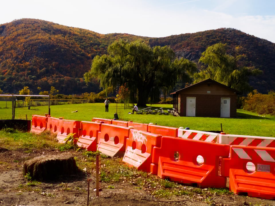 A park along the hudson. Orange construction barriers are visible in the foreground, with hills in the background.