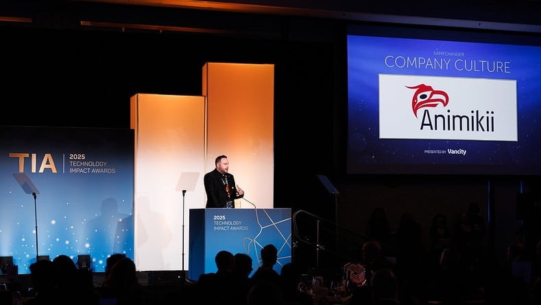 A man stands at a podium on stage in a dimly lit ballroom. There is a screen on the right that has the projection of a company logo for Animikii Indigenous Technology, and says Winner - Company Culture Award