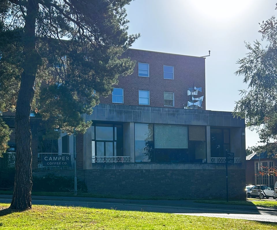 A concrete and brick building on a slope. The single storey concrete block forms a foyer, with large plate glass windows, that rests on a rubblestone wall. The brick building rises behind it, and is a simple block with plain windows.
