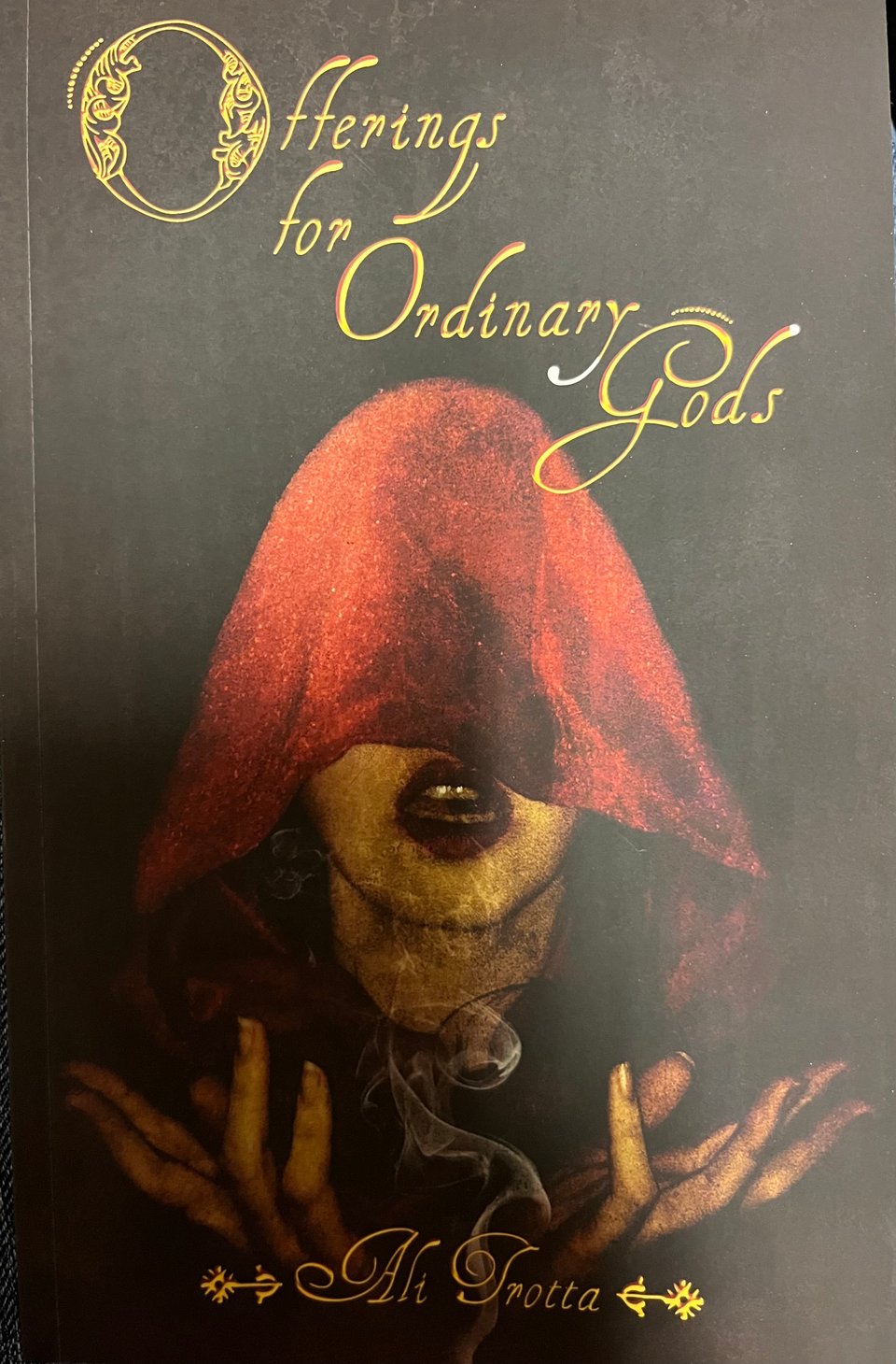 The cover of my poetry collection, in which a woman wearing a red cloak is holding up her hands, with smoke between them. The title (Offerings for Ordinary Gods) and the author name (Ali Trotta) are rendered in beautiful gold script