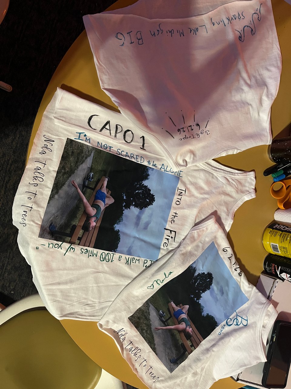 Three Tightrope tank tops on a mustard yellow table. Two show the back of the bank with pictures of Nola lying on a bench, the other shows the front of the tank with the text "Tightrope" on the upper right side. All three are decorated with lyrics from the song, doodles, the song release date, and signatures from Nola and the gremlins.