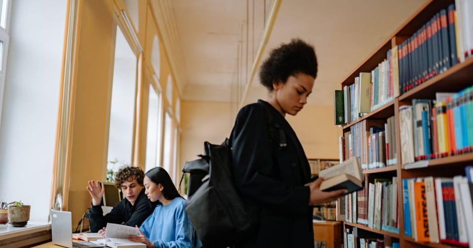Image of a Black student browsing library shelves, with student using a computer behind them.