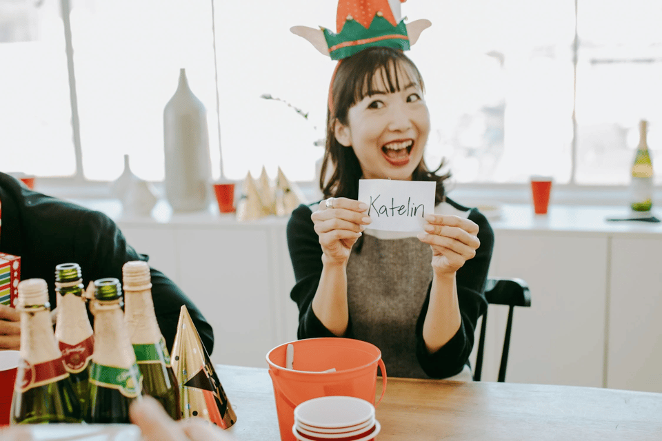 A photo of a woman sitting at a table at what appears to be a party — there were a few bottles of champagne on the table, and she is wearing a party hat. She is holding up a White piece of paper with the name "Katelin", and she has a huge open-mouthed smile.