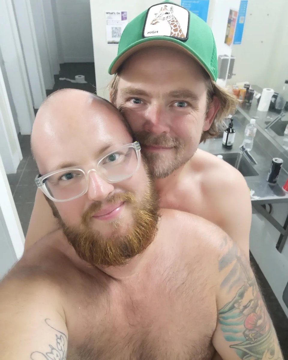 This is a selfie of two smiling, shirtless men — the author and their friend Charlie — embracing in a bathroom setting at a festival. The man in the front has a beard, wears clear-framed glasses, and has a colorful tattoo on his arm. The man behind him is wearing a green cap with a giraffe graphic and the word “HIGH” on it. They appear to be happy and close, with the background showing a row of sinks and toiletries.