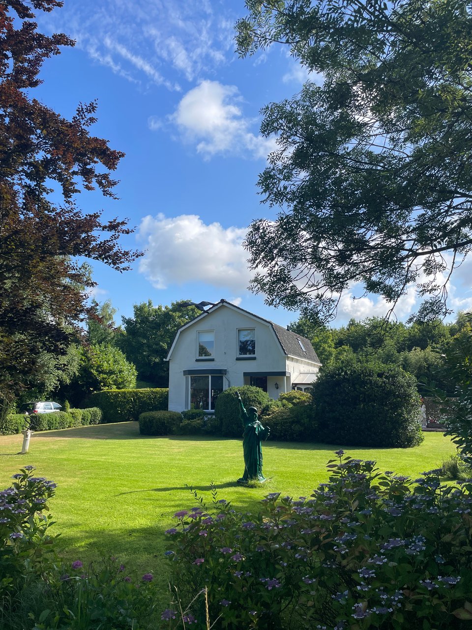 A small version of the statue of liberty stands in the middle of a lawn in front of a garden surrounded by trees. In the background is a two story farm house.