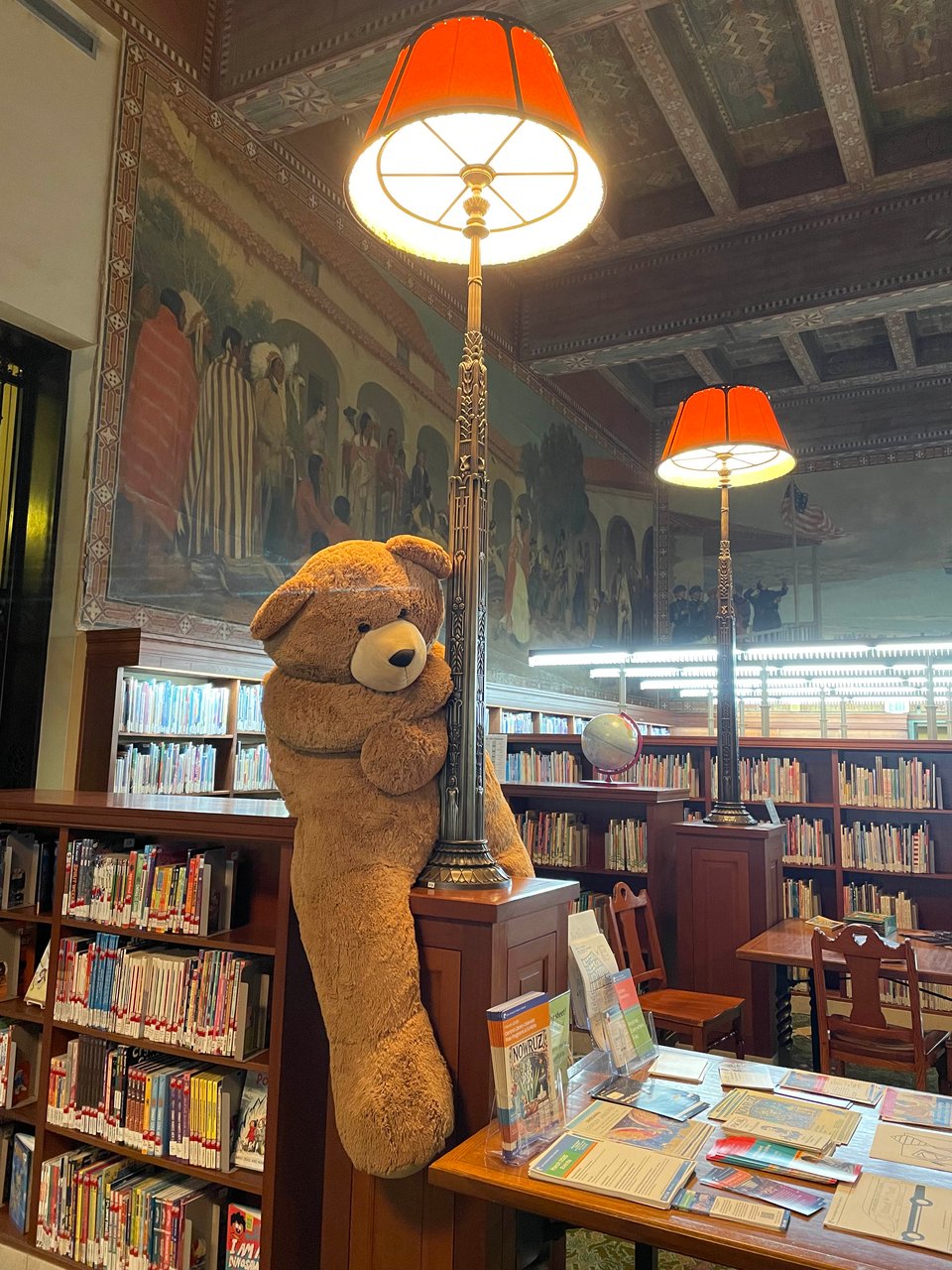A large stuffed bear leans wistfully on a tall, ornate lamp in a room full of dark wood and books