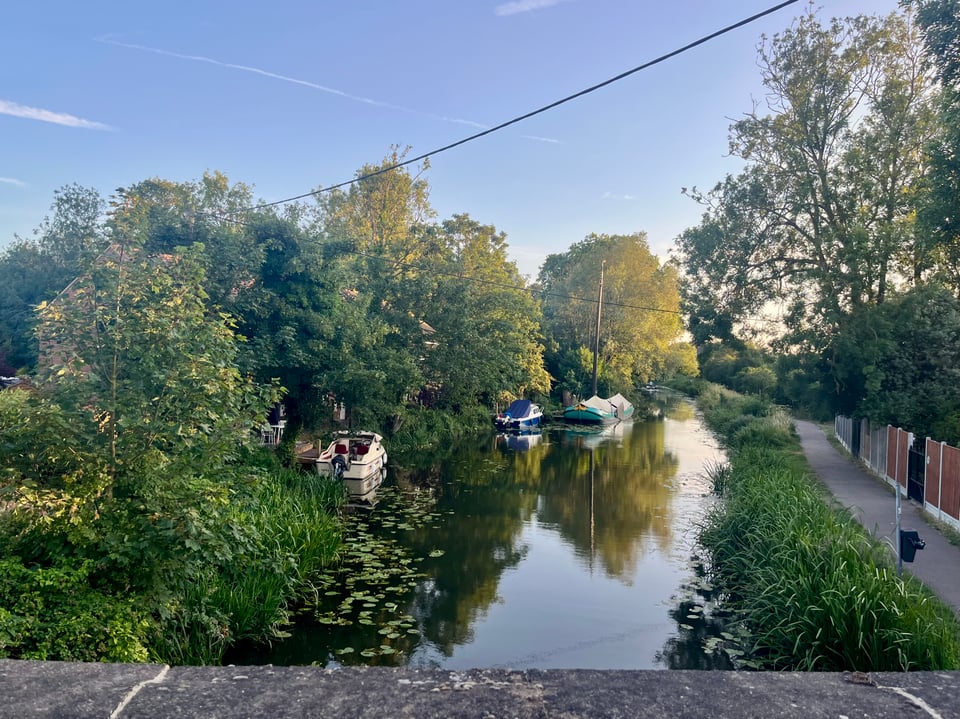 A river with trees and greenery down one side, and a path and grasses on the other. The river disappears into the horizon, with a few boats moored on one side. The sky is a light blue with wisps of cloud, and the sun is setting in the distance.