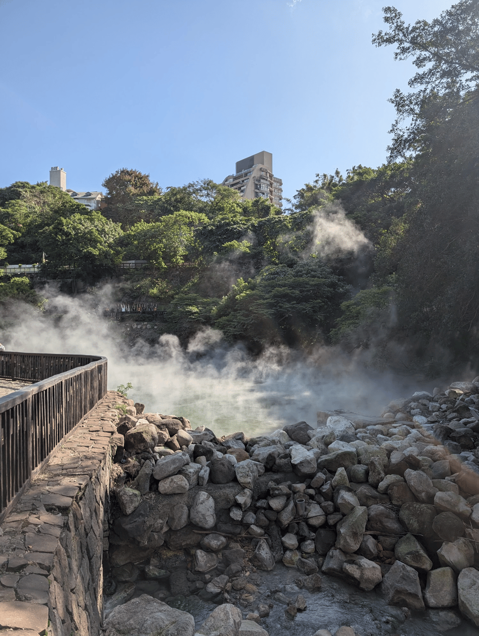 A pile of rocks is in the foreground damming a body of greenish water that's emitting a thick misty steam. behind it are trees and a hotel in the distance. On the side is wooden railing and a walkway.