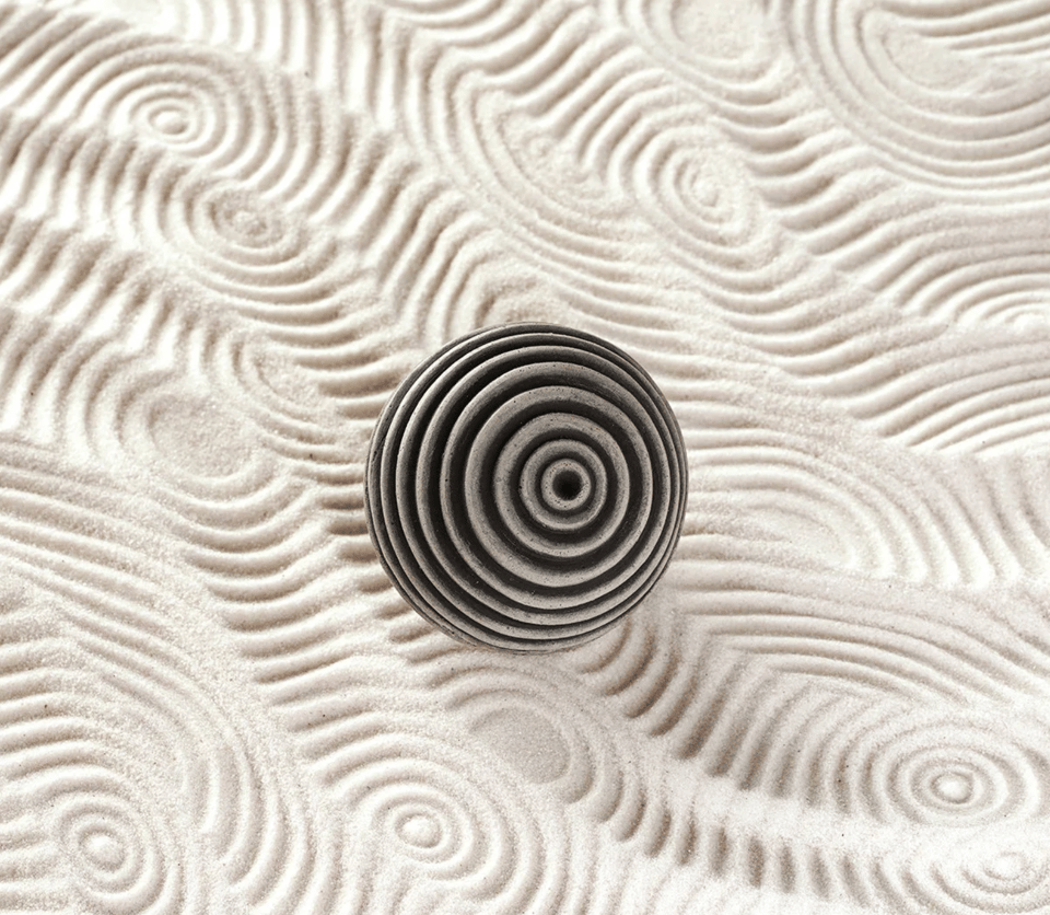 A photo of a circular ball, concrete-colored, with a pattern of circular ridges in concentric circles. It is on top of sand, and in all directions the sand is imprinted with curving, undulating patterns from the ball rolling around