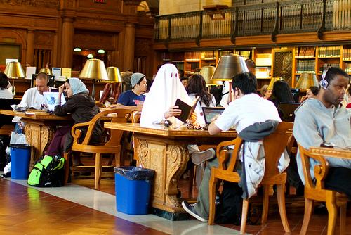 A classic white-sheet ghost sits reading in a crowded NYPL.