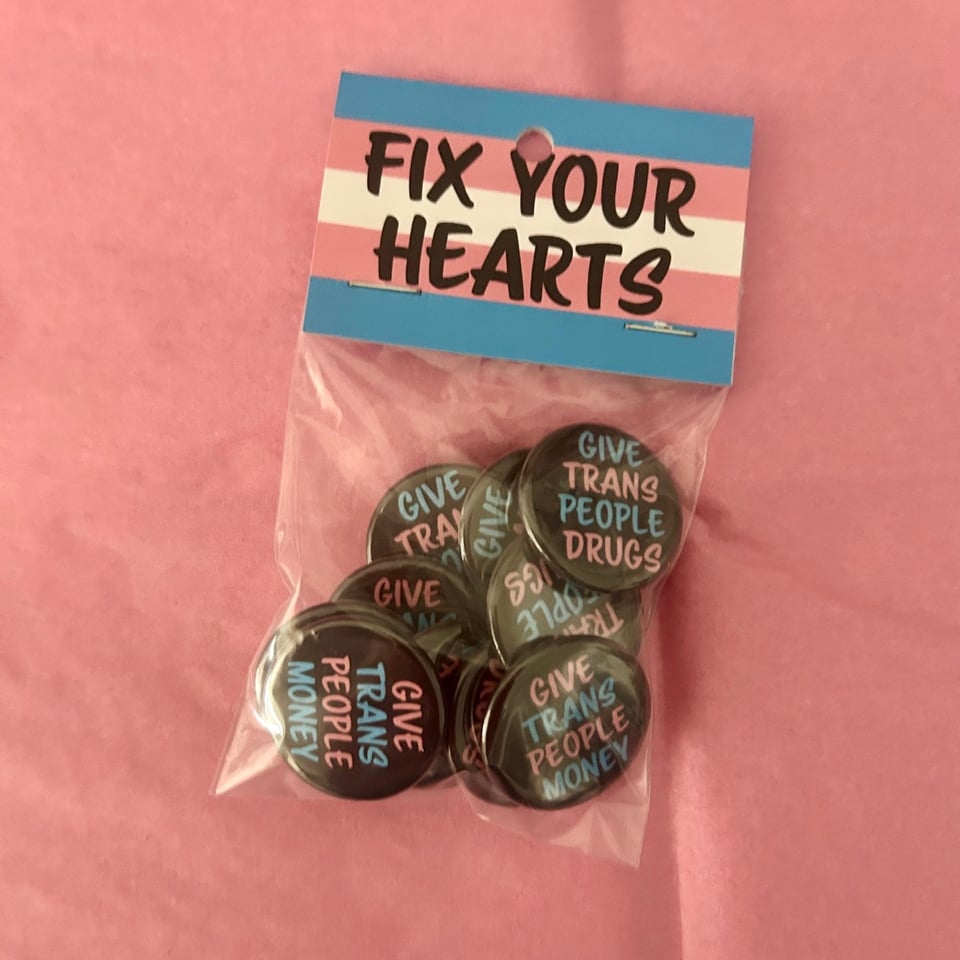 A bag of pins that say GIVE TRANS PEOPLE MONEY and GIVE TRANS PEOPLE DRUGS. The label on the pins says FIX YOUR HEARTS.