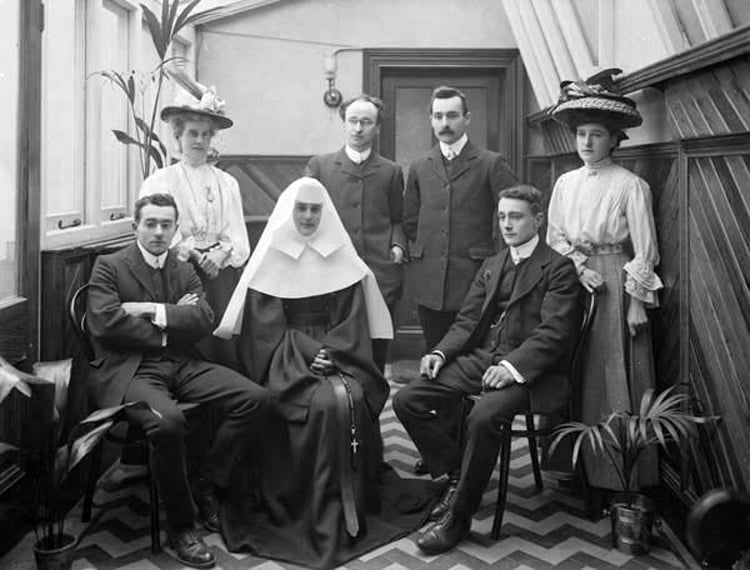 A nun seeks mercy, surrounded by some well dressed people, one woman has the most extravagant hat