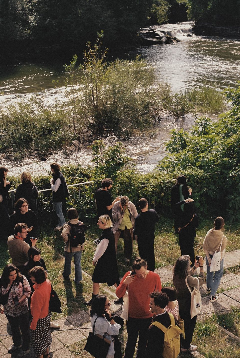 [People cluster a meander around a green area next to a river, holding their phones to their ears.]