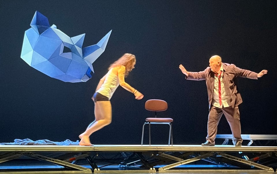 A dancer and an actor perform on stage, with a large, stylized blue rhinoceros head floating above them and a single chair positioned between them.