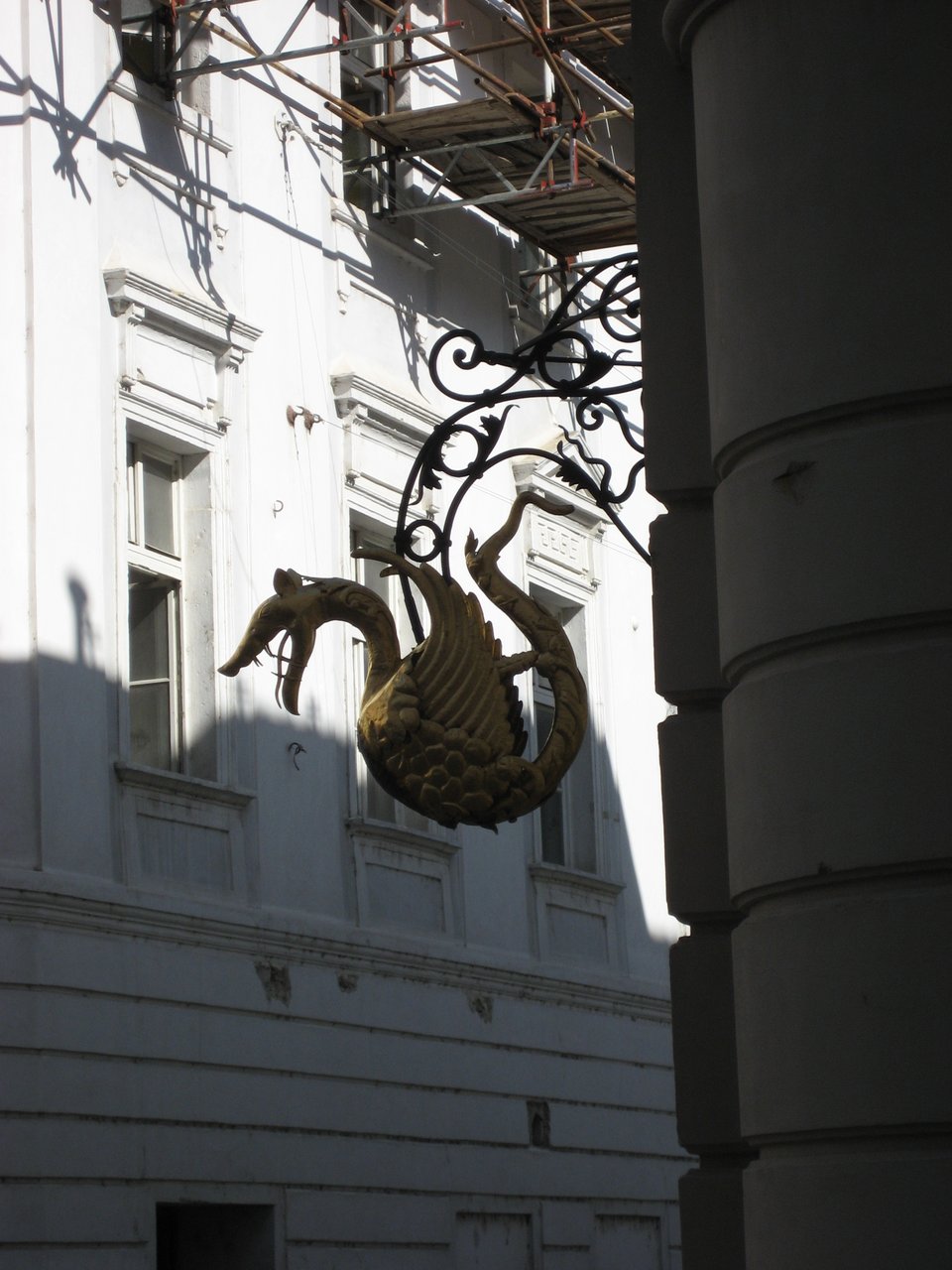 A sign in the form of a dragon with spread wings and an open mouth, hanging from a building. Behind it is a white wall with windows.