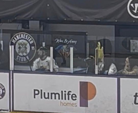 A player dressed in white sitting in a plexiglass booth / box. Man with yellow foam head standing to one side, staring at him.