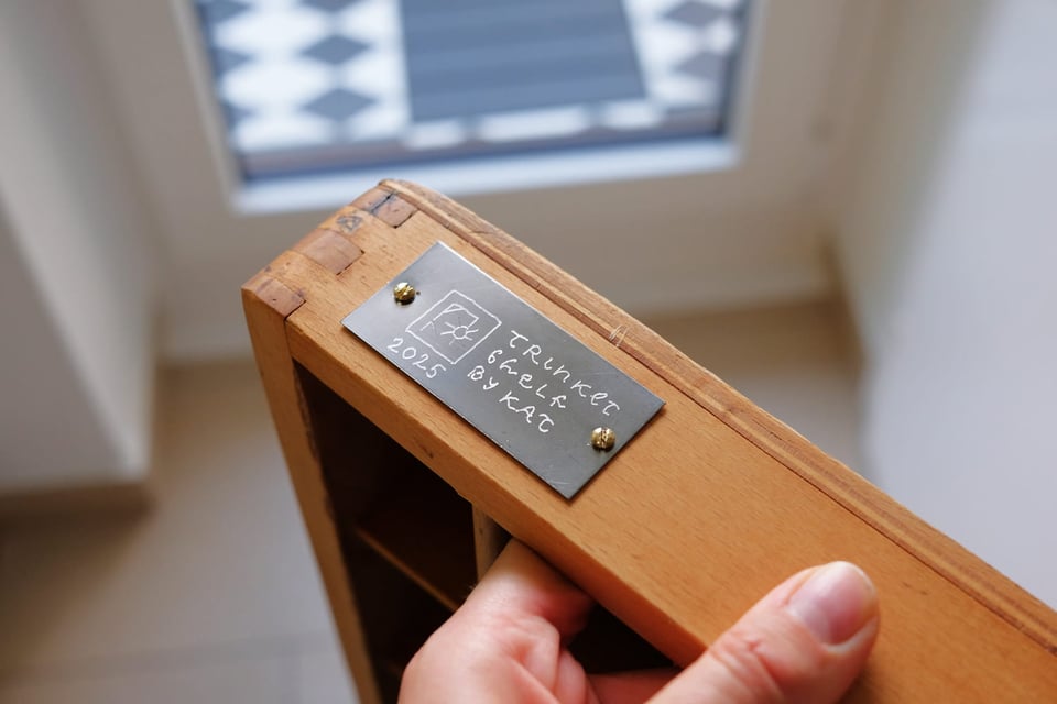 A photo of my hand holding up a corner of the trinket shelf. A metal plaque has been screwed onto it. The plaque says "Trinket Shelf by Kat" next to a logo of a hanged sun and the year 2025.