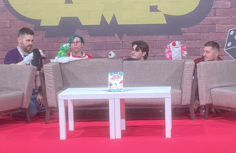 Four people crouch behind couches at a convention room, partway through an actual play performance. Each person holds a puppet. In front, Sock Puppets sits on a stand.