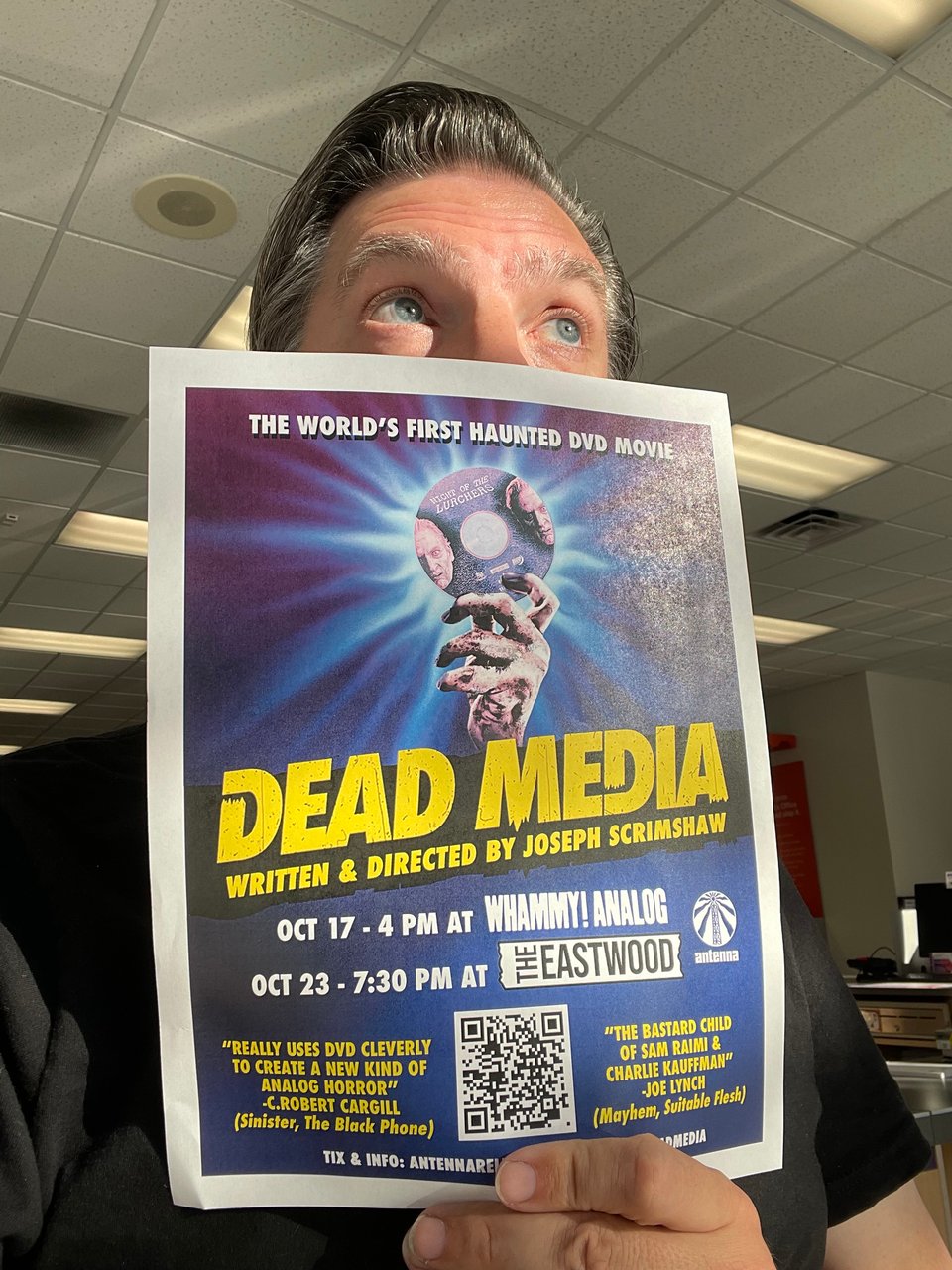 A man holds a paper ad for the horror film Dead Media in front of his face. The man, for better or worse, is in a Fed-Ex Office