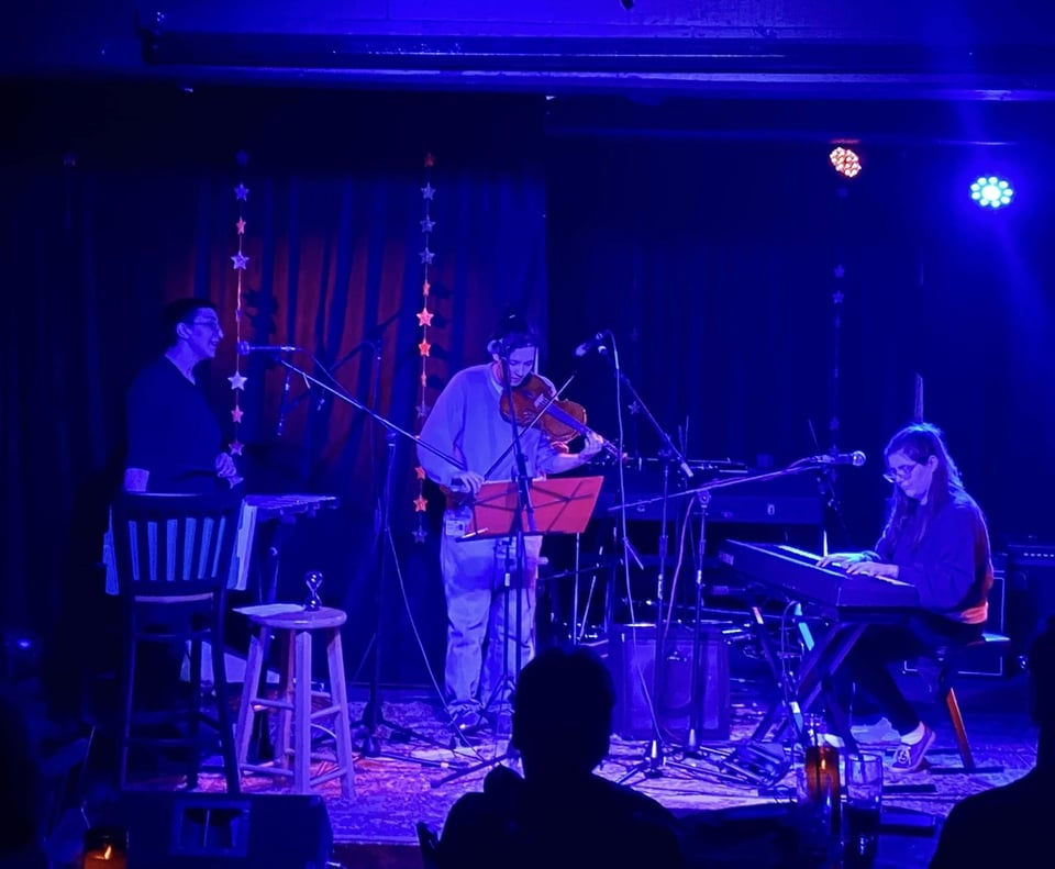 Three musicians play percussion, violin and keyboards on a stage bathed in blue light