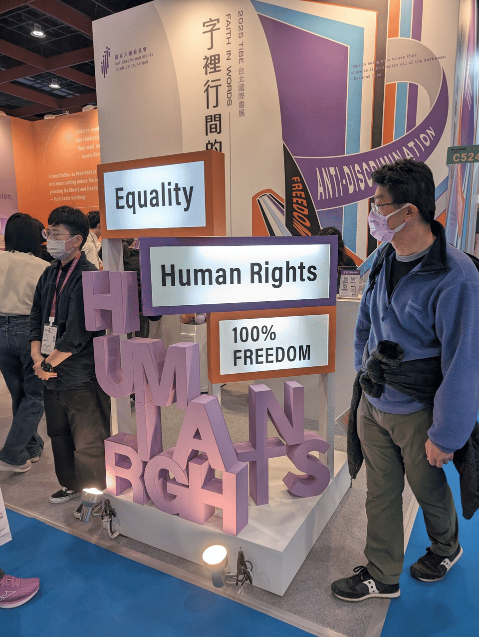A display for the Human Rights Commission, which says "Faith in Words," "Equality," "Human Rights," and "100% Freedom." One person is standing next to it looking toward the crowd. Another person is standing on the other side, looking at the display. In the background you can see a graphic that says "Anti-discrimination" and "freedom" alongside illustrations of books.