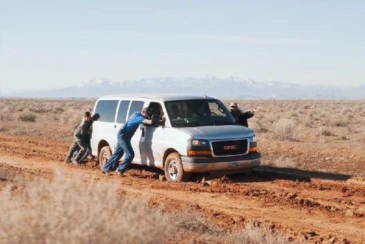 a group of people pushing a stocked van on a dirt road