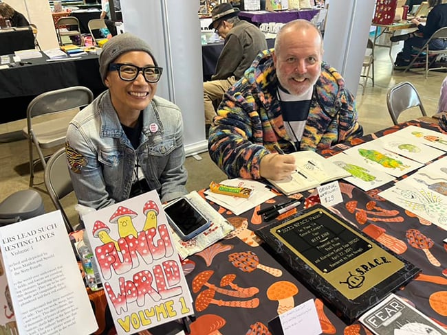 Ione is on the left, Matt is on the right. They are sitting behind a table; there are comics and pieces of art on the table.