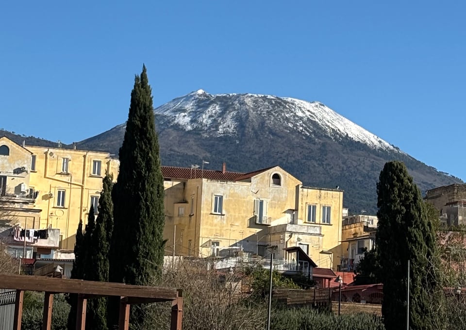 The volcano Vesuvius with snow on it, buildings and trees in front, very blue sky above.