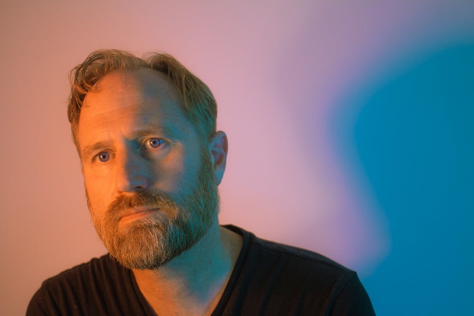 A man with short hair parted on the left and a beard looks just past the camera