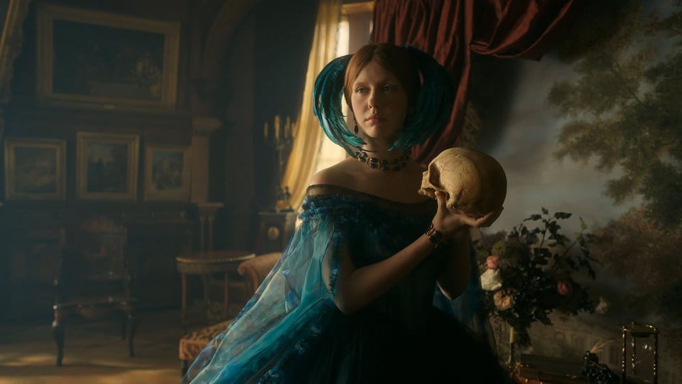 A white woman wearing a blue dress and headpiece, holding a skull as she looks left off-screen.