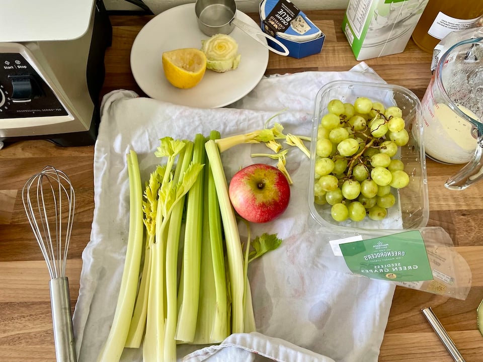 Celery, apple, grapes, a whisk and other cooking components on a counter