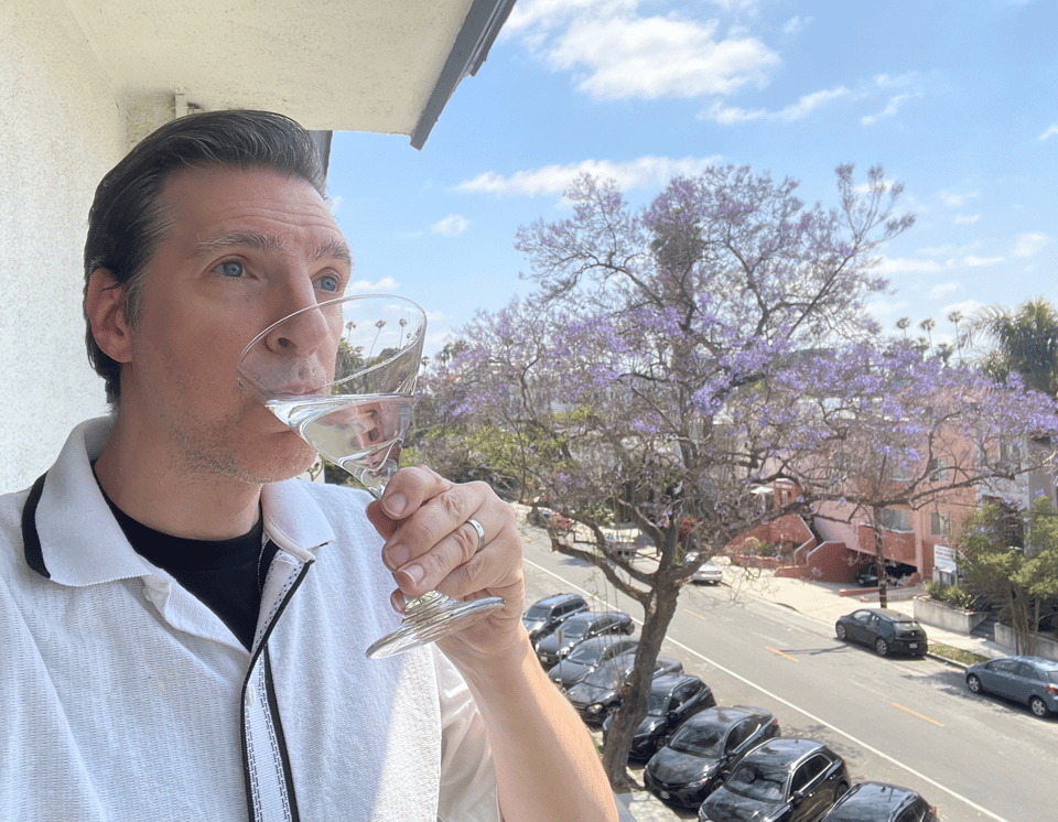 A man in a white and black 60s era swing shirt sips a fake martini on a balcony
