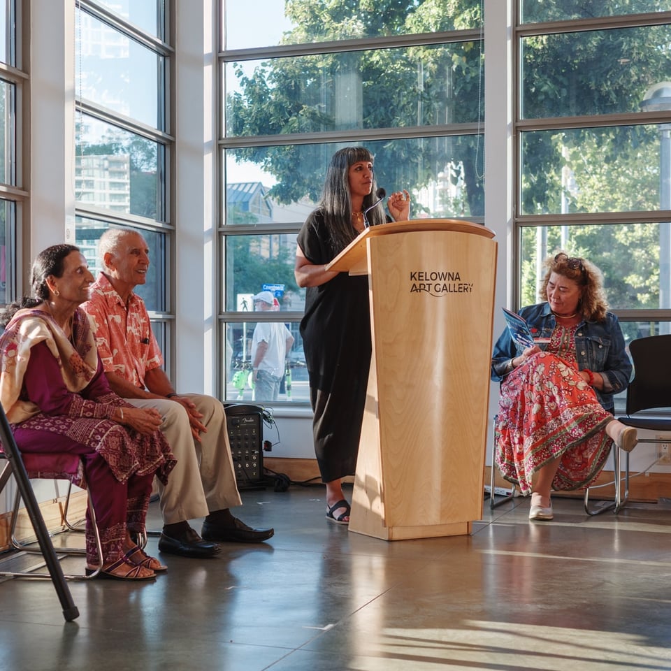 A woman with long dark hair and bangs speaks at a wooden podium marked 'Kelowna Art Gallery' in a bright, modern gallery space with floor-to-ceiling windows. Three people sit in chairs listening - two older adults on the left (one in a purple patterned garment, one in a coral-colored shirt) and a woman in a denim jacket and floral dress on the right. Sunlight streams through the large windows, showing green trees and urban buildings outside.