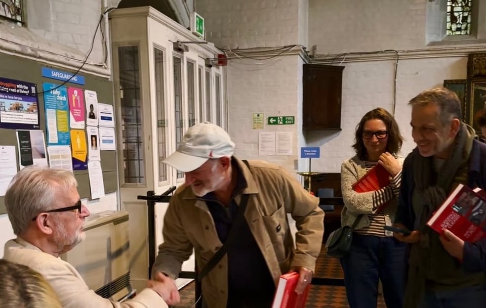 A man seated at a table shakes another mans hand as he is holding a book to have signed. Two other people holding books look on smiling.