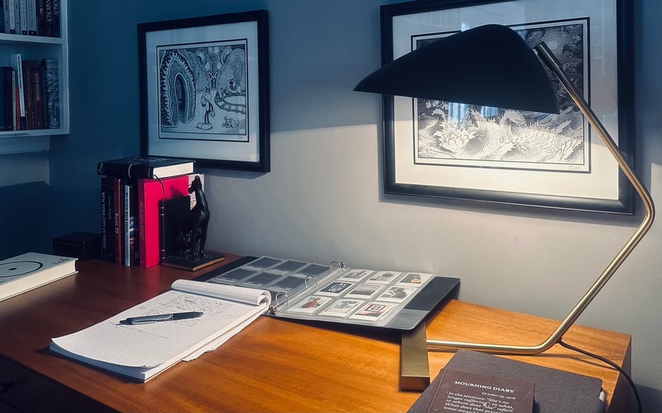 photo of desk with mid-century lamp, books, a binder filled with Instax photos, and a notebook and pen. Some Jim Woodring prints are on the wall behind it.