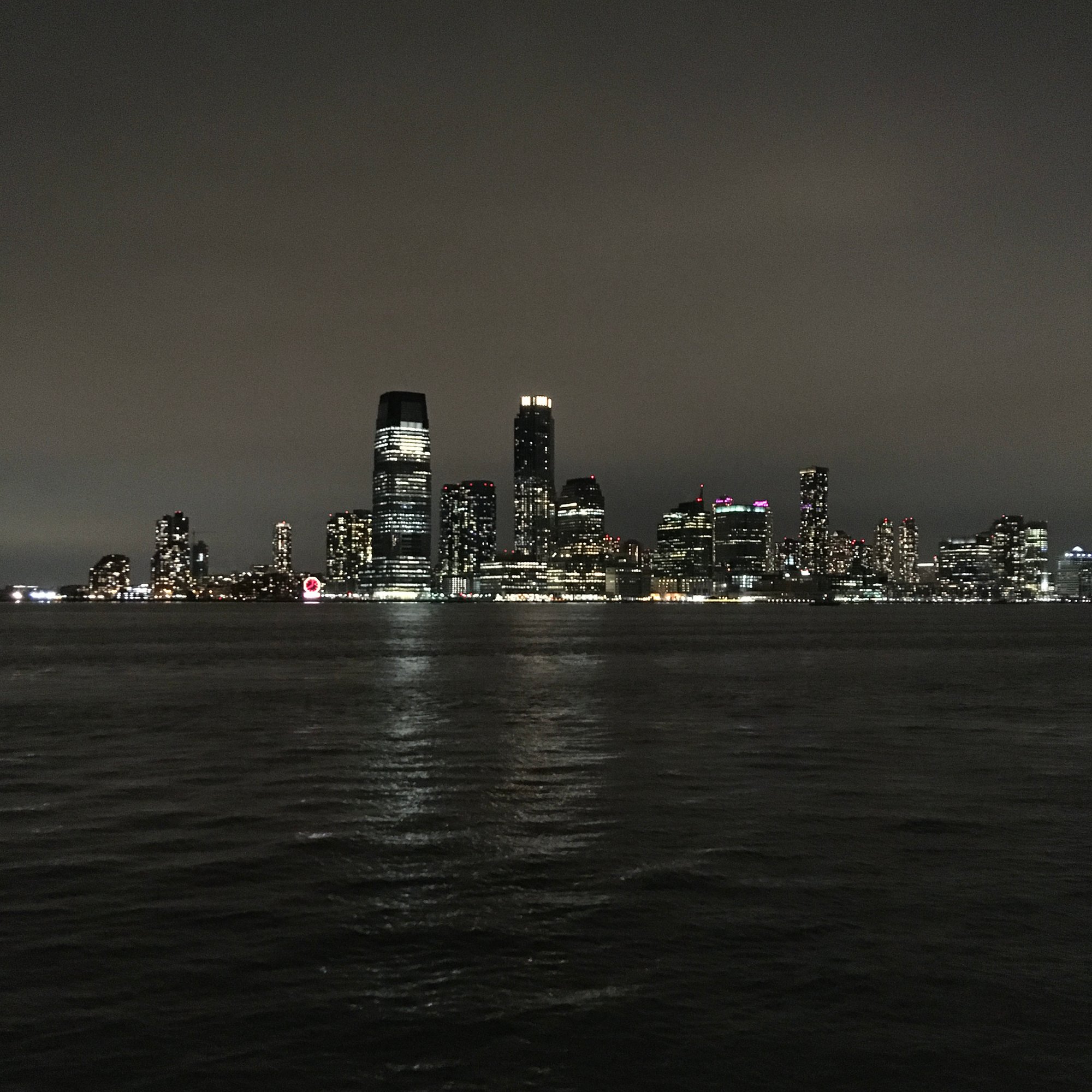 Photo of the downtown New Jersey skyline at night, from across the Hudson River. The water and sky are dark grey, clouded, illuminated only by the light of buildings and skyscrapers.