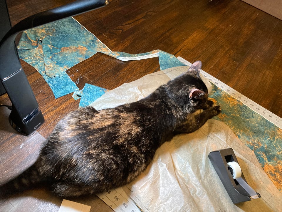 A tortoiseshell cat is sitting on fabric and a paper pattern under an ottlite, clearly they are being cutely in the way