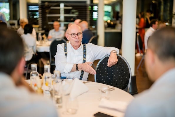 Photo of a man at a large round table talking with animation to the people nearest us, who are out of focus.