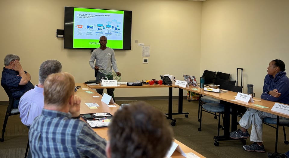 Shantae presents to a room of NRG members with a presentation displayed on a wall-mounted screen behind him.