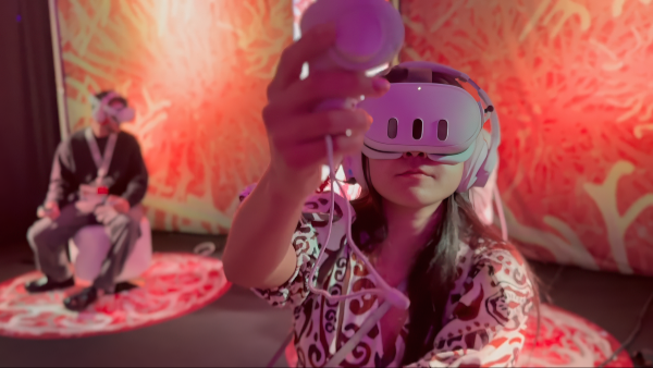 A woman wearing a VR headset and headphones interacts with a controller in a vibrant, immersive installation with abstract, organic visuals on the walls. In the background, another person is also wearing a VR headset while seated on a stool. The scene is lit with pink and red tones, creating a surreal and engaging atmosphere.