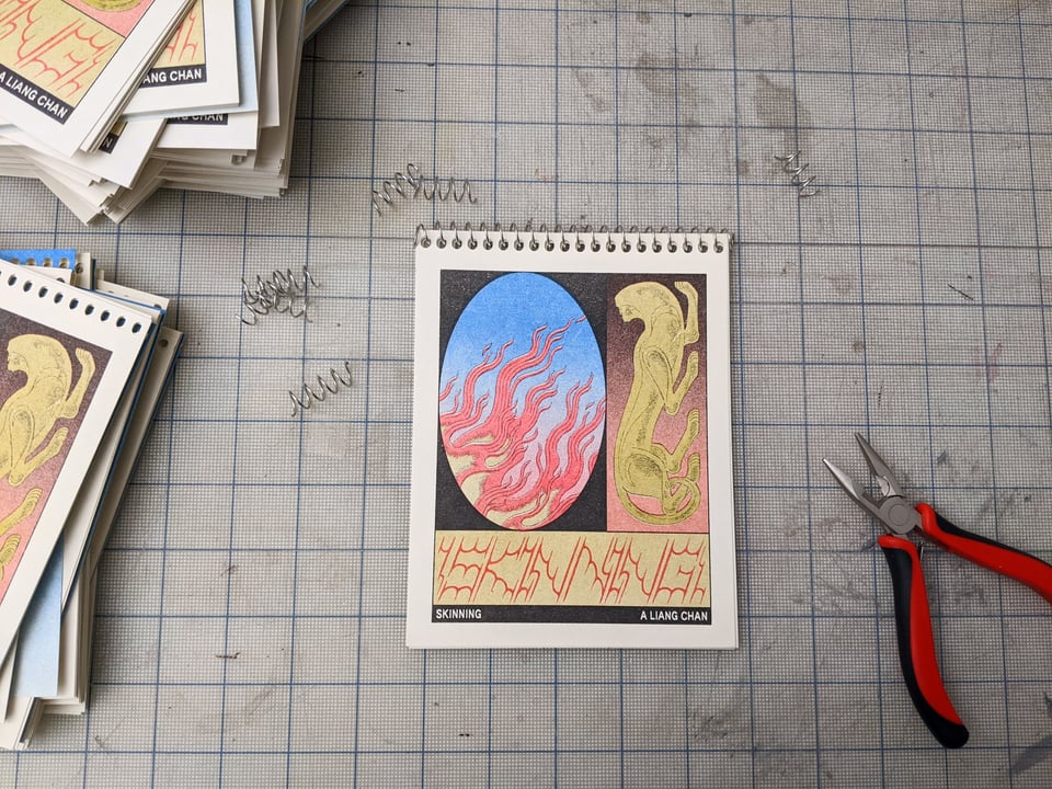 a riso printed spiral bound book sits on a cutting mat, trimmed metal coils around it. the cover features fire against a blue sky, a figurine of an animal missing its head, and the title "skinning" in spiky red lettering.