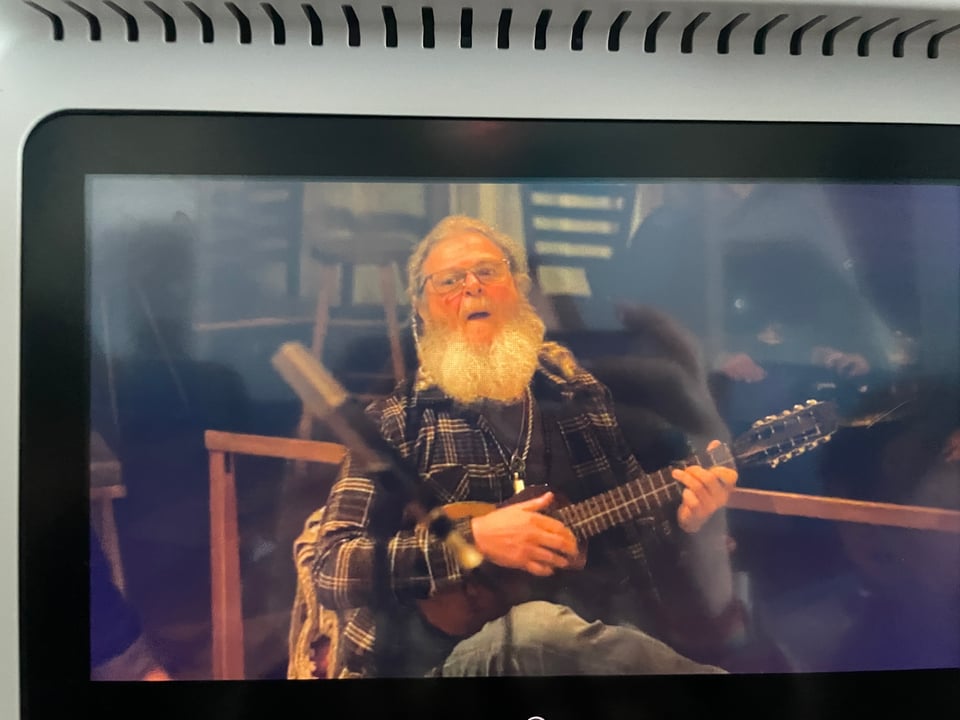 A bearded man with an interesting small guitar dressed in a flannel shirt plays at a party.